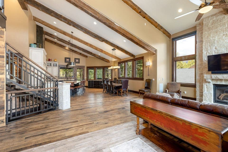 Spacious open-concept living area with wooden beams, hardwood floors, a dining table, a staircase with metal railing, modern kitchen, large windows, a fireplace, and a ceiling fan.