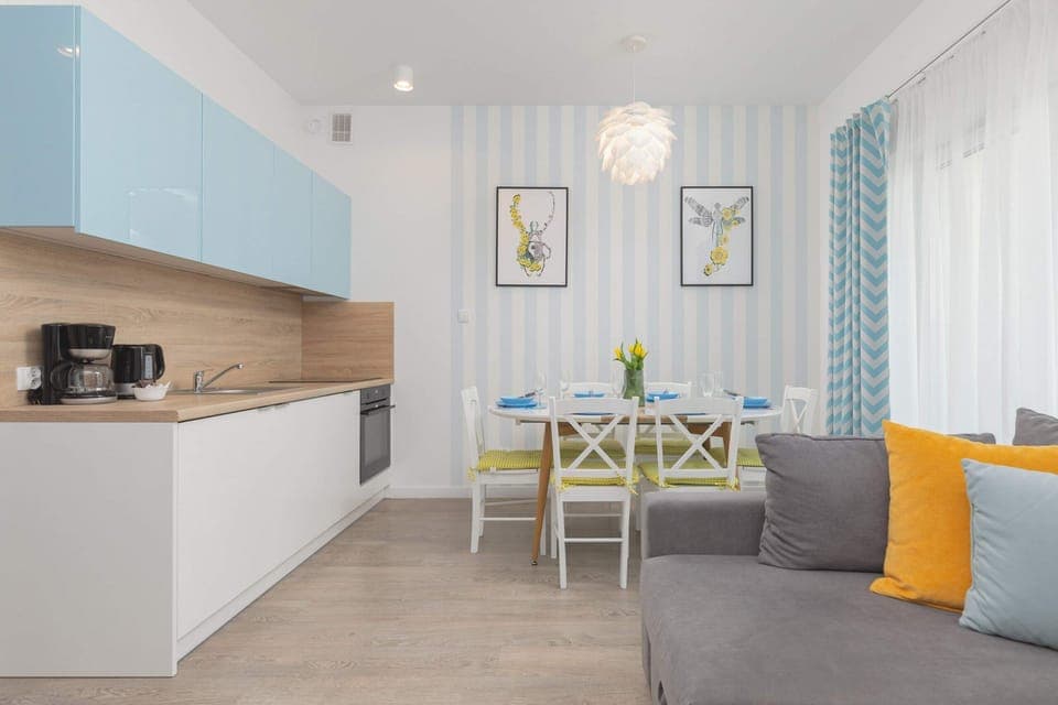A well-lit, open-plan kitchen connected to a dining space with a wooden table and colorful accents. The bright interior feels fresh and modern.
