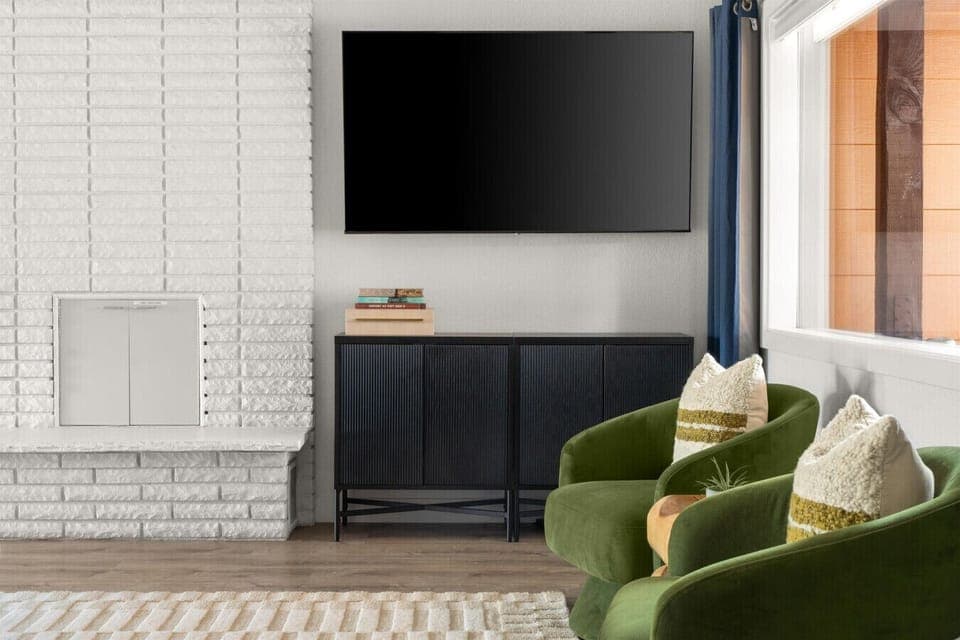 Unwind in this cozy nook with plush green chairs, a sleek smart TV, and a charming white brick fireplace—perfect for movie nights or quiet mornings with a view of the Arizona sunshine just outside the window.