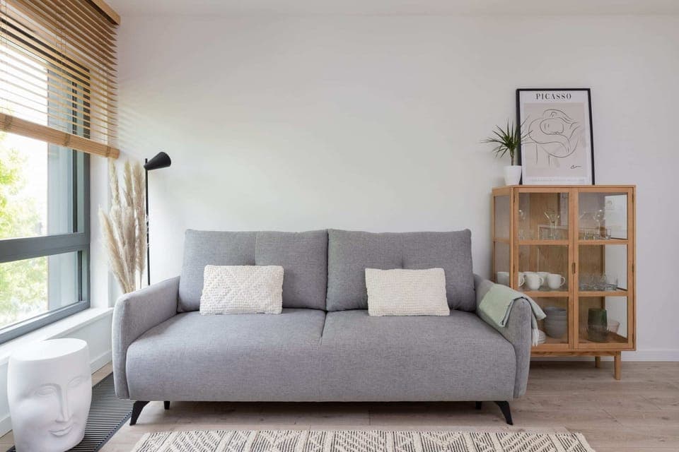 A cozy living room with a gray sofa, a wooden side table, and a minimalistic bookshelf, complemented by soft natural light from large windows.