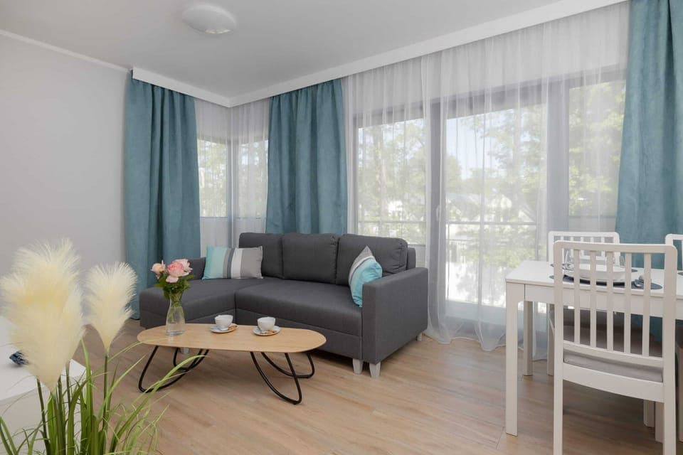 A bright living room with a blue sofa, large windows, and sheer curtains. A round coffee table sits in the center, surrounded by modern decor.