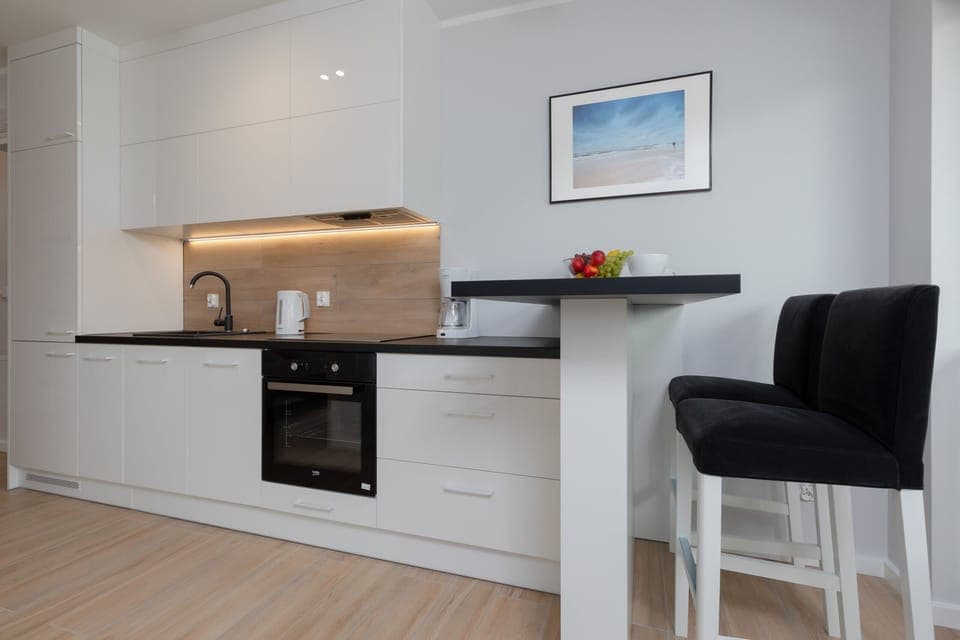 A sleek and minimalist kitchen has white cabinetry, wooden accents, and a black countertop with a barstool tucked in.