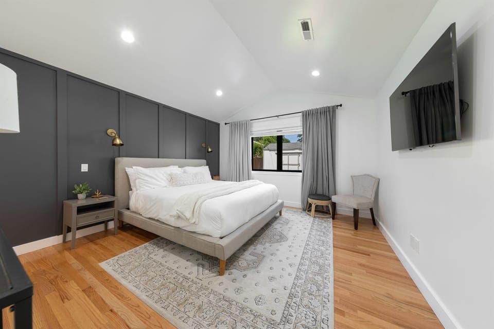 Spacious ensuite bedroom featuring a luxurious California King hybrid mattress from Saatva, ambient sconce lighting, a 55-inch TV, and large windows overlooking the backyard. Blackout curtains make it perfect for sleeping in and fully enjoying the comfort