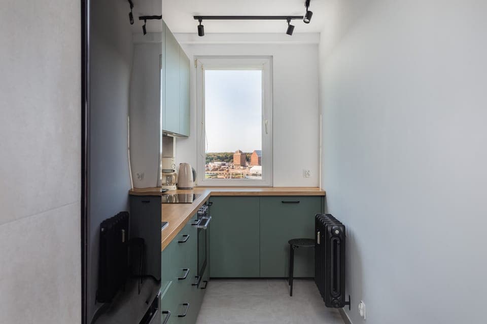 A compact kitchen with sage green cabinets, a large window, and a minimalist style.
