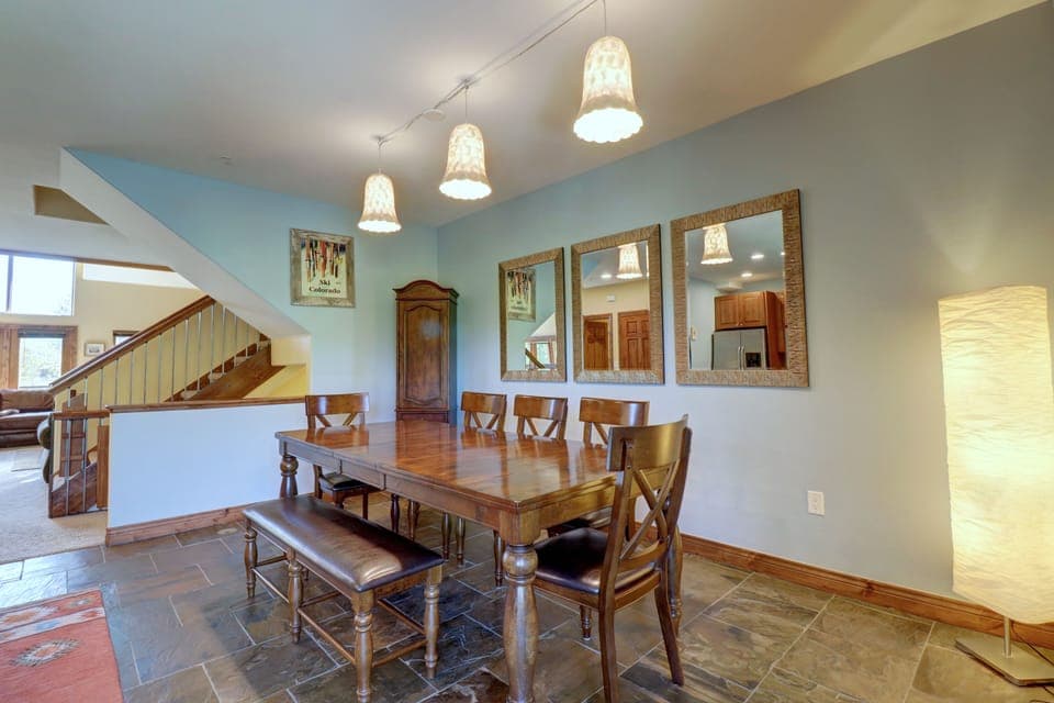 A dining room with a wooden table, six chairs, and a bench. Three pendant lights hang above. The walls feature three mirrors and two artworks, with a wooden cabinet in the background.