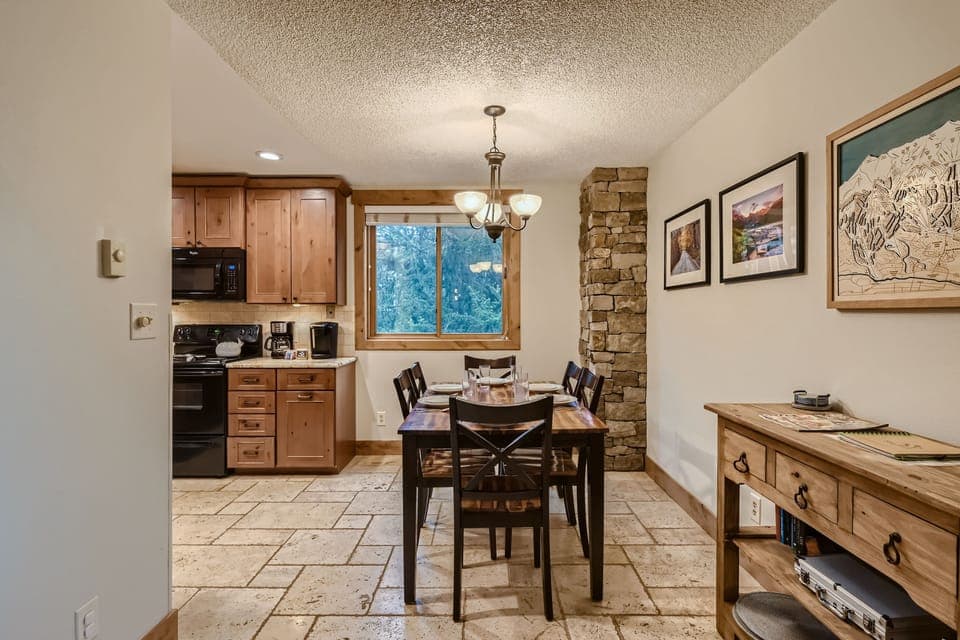 A small dining area with a wooden table and four chairs, adjacent to a kitchen. The space has stone tile flooring, a window, and decorated walls. A chandelier hangs above the table.