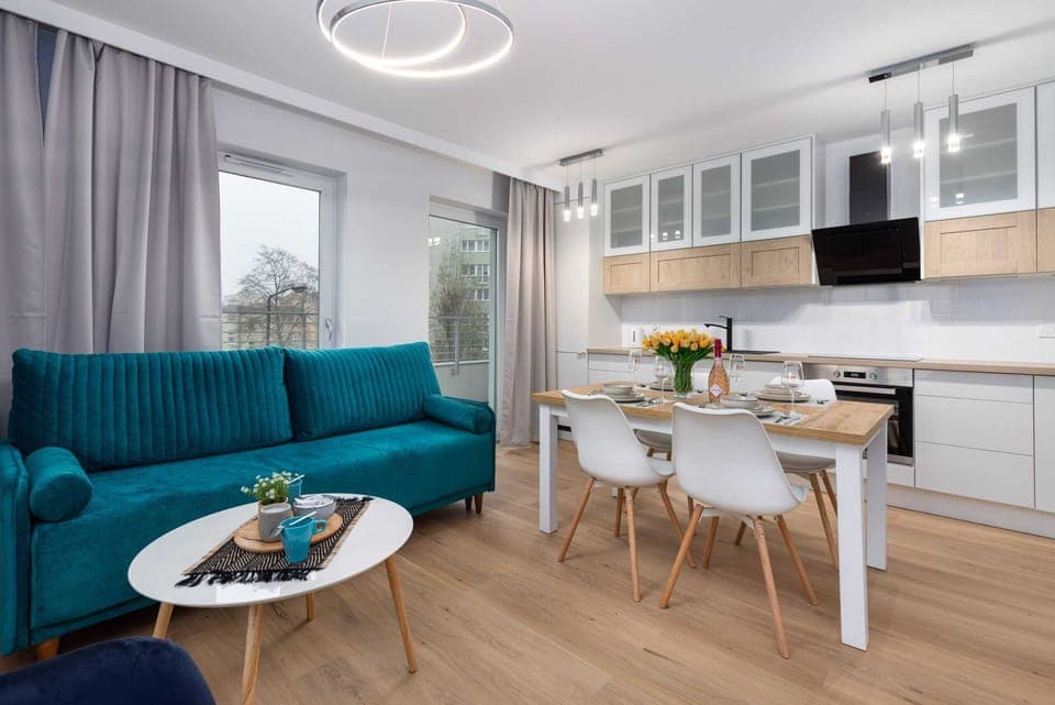 A cozy living room with a stylish green sofa, a small coffee table, and a dining table for four, illuminated by natural light.
