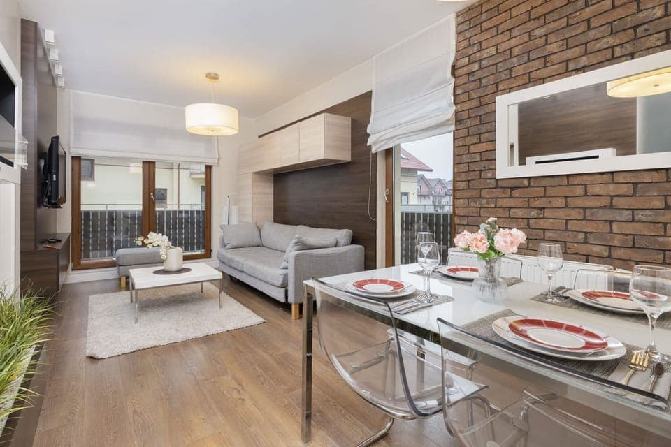 A bright living room with gray and neutral-toned furniture, including a sofa and a small coffee table. Large windows allow plenty of natural light to fill the space.