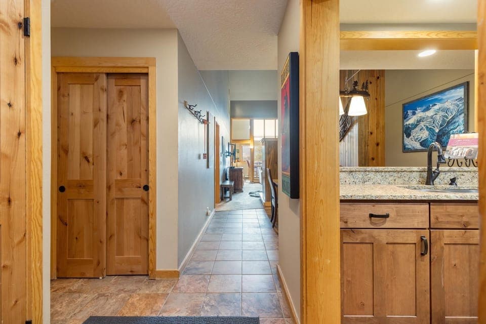 Hallway in a home with wooden doors on the left, a tiled floor, and a kitchen area with a granite countertop on the right. The hallway leads to a well-lit living space.