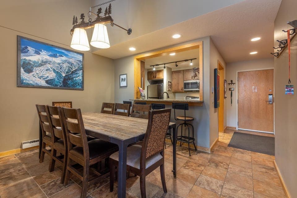 A dining room with a wooden table and six chairs is adjacent to a kitchen with an open counter, two bar stools, and modern appliances. A painting and pendant lights decorate the space.