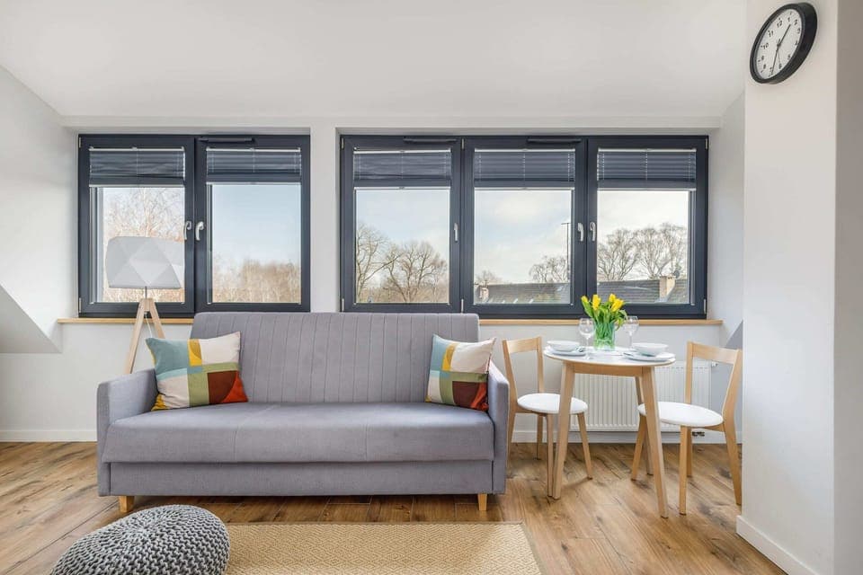 A cozy living room with a gray sofa, colorful cushions, and a dining table with wooden chairs, complemented by large windows allowing ample natural light.