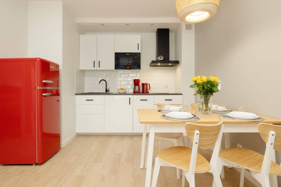 A modern kitchen and dining area featuring a red fridge, white cabinetry, and a wooden dining table set for a meal.
