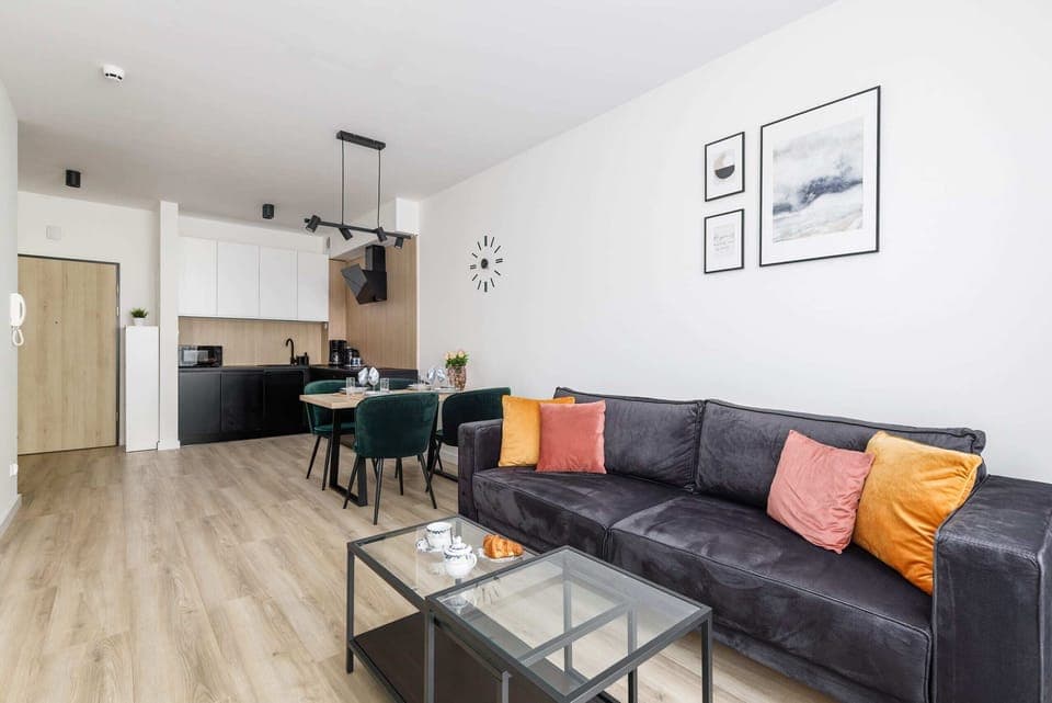 A bright living room with a black sofa, decorated with vibrant cushions, a coffee table, and a large window.