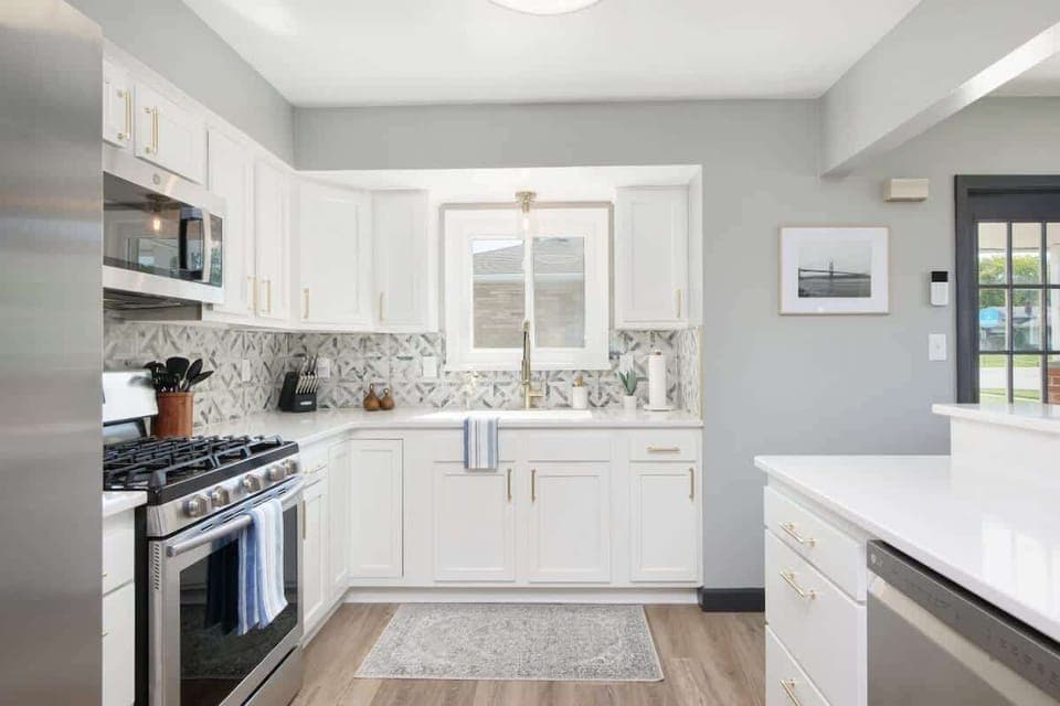 - Modern kitchen with stainless steel appliances and gas range   - White cabinets with gold handles and geometric tile backsplash   - Bright space with window above sink for a home style feel  