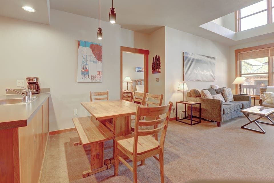 Step into your bright, open living space where natural light flows through skylights, warming the cozy dining area and comfortable seating. You'll love gathering around the rustic wooden table while enjoying mountain views through large windows.