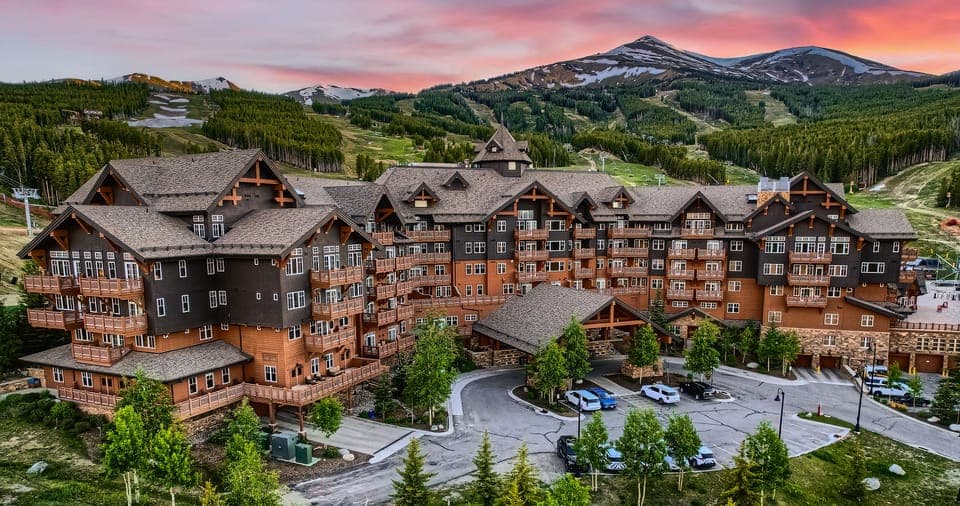 A large rustic mountain lodge with multiple gables and balconies, surrounded by lush greenery and set against a backdrop of forested hills and a pink-hued sunset sky.