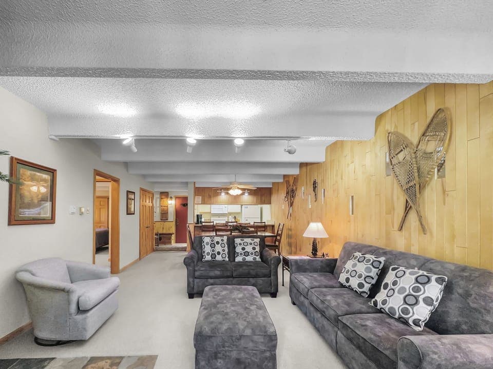 A living room with two couches, an armchair, a coffee table, and framed pictures. The room has wooden paneling, snowshoes as décor, and an open view to the adjacent kitchen and dining area.