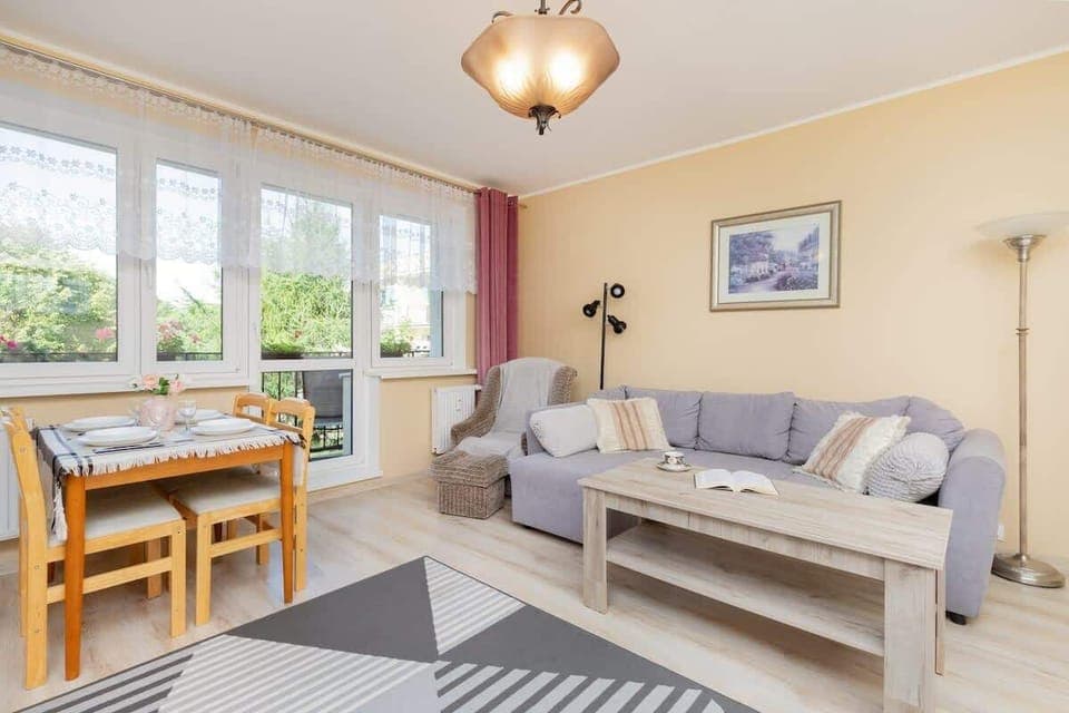 A bright living room with a dining table, gray sofa, and a large window, creating a cozy and inviting space.

