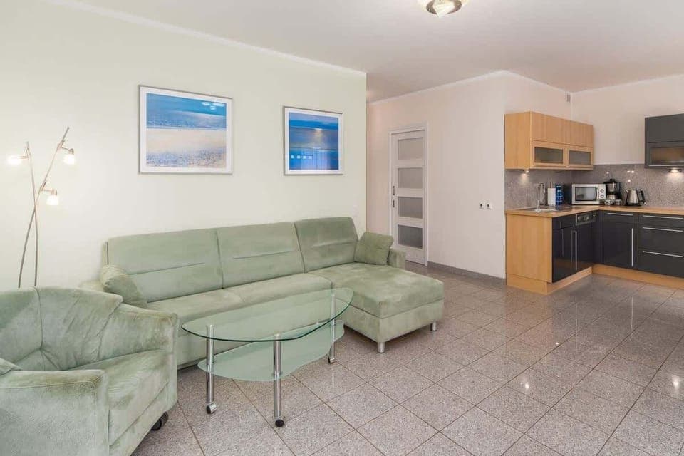 A spacious living room with a green sectional sofa, a coffee table, and an open-plan connection to the kitchen.
