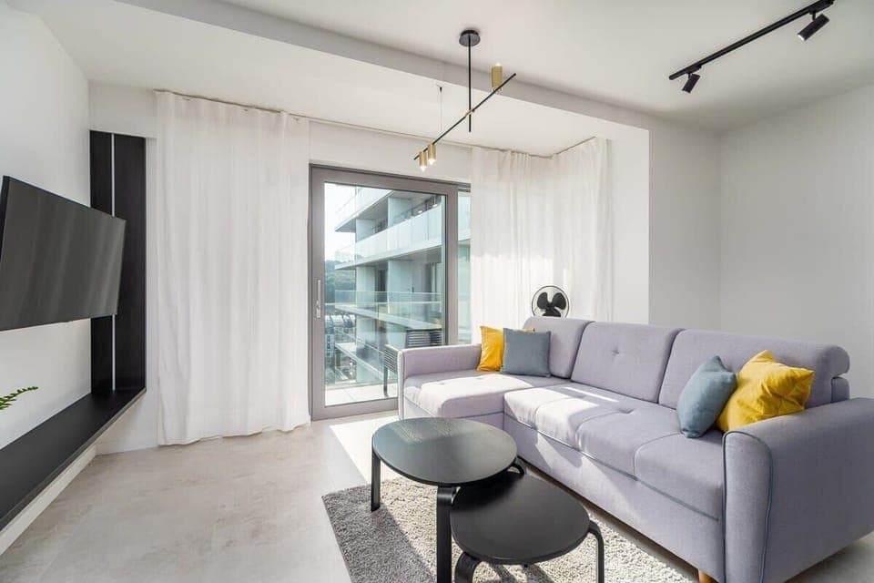 A bright living room with a gray sofa, a round coffee table, and a wall-mounted TV. Sheer curtains let in natural light.
