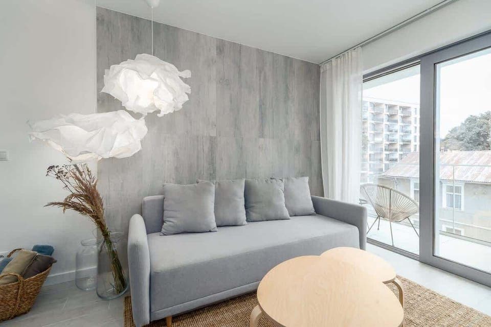 A bright living room with a gray sofa, round coffee table, and floor-to-ceiling windows opening onto a balcony.
