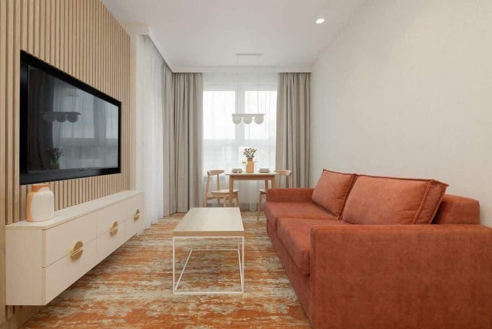 A cozy living room with a large wall-mounted TV, an orange sofa, and a patterned rug, creating a warm and inviting atmosphere.
