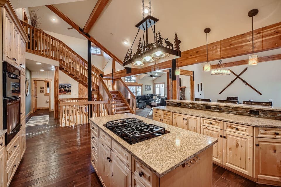 A spacious kitchen with wooden cabinets, a large island with a cooktop, and a rustic chandelier. In the background, there's a wooden staircase and a living area with large windows.